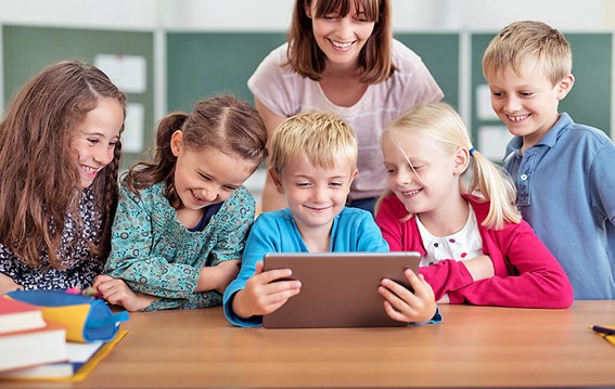 Criança segura o tablet na sala de aula enquanto a professora e outros alunos observam o conteúdo.