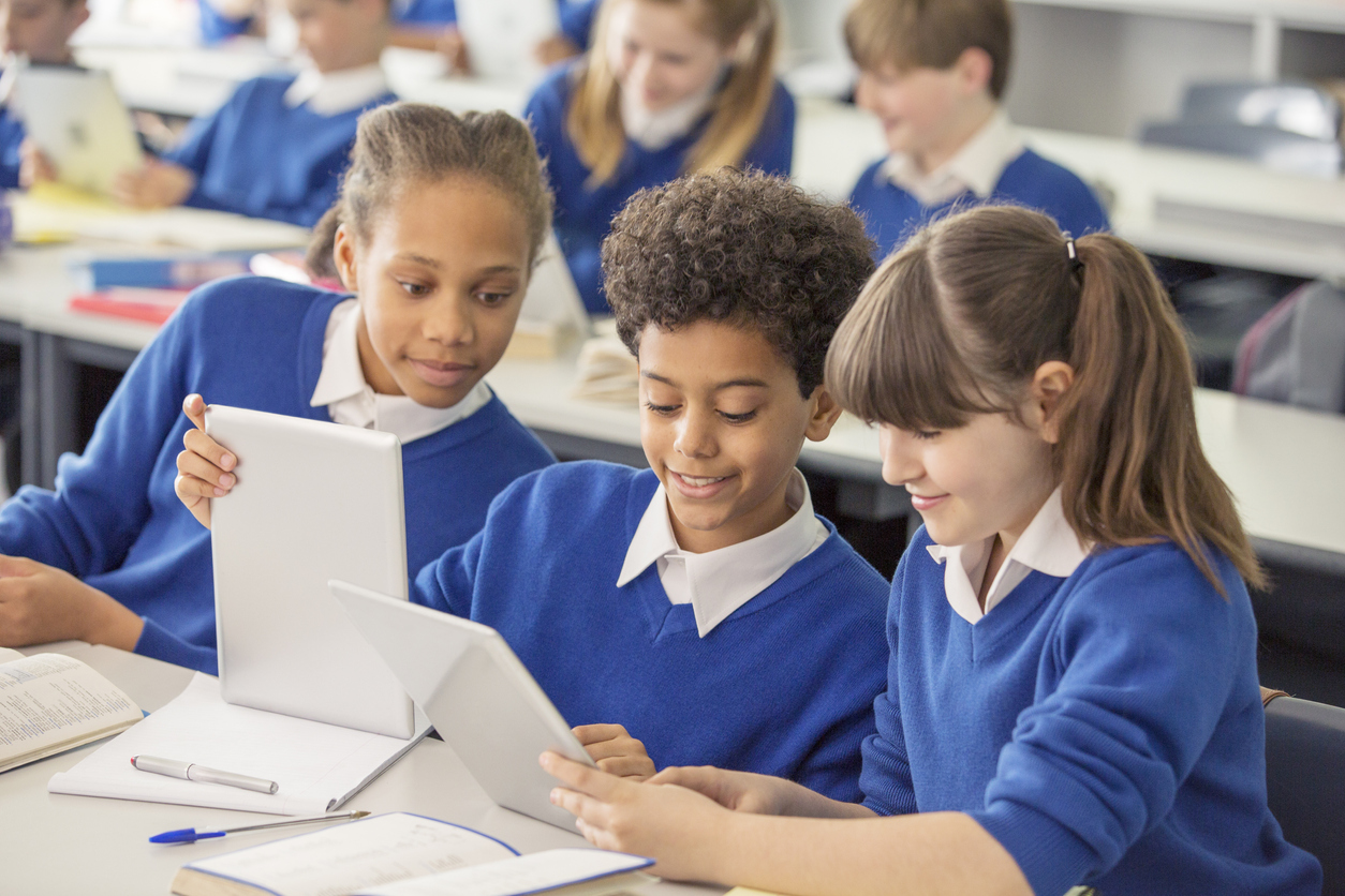 Crianças do ensino fundamental usando uniformes escolares azuis usando tablets digitais na mesa em sala de aula