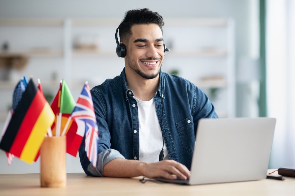 Um jovem sorridente usando fones de ouvido e digitando no notebook. Há bandeiras de diferentes países em cima da mesa.