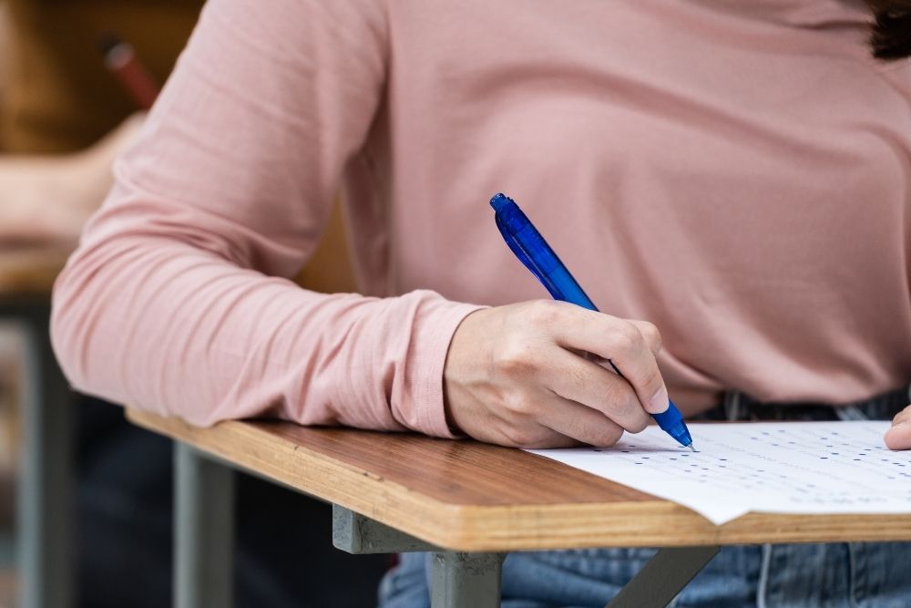 Pessoa adulta escrevendo em um teste, usando uma caneta azul e uma blusa rosa. A imagem foca nas mãos e na atividade de escrita.