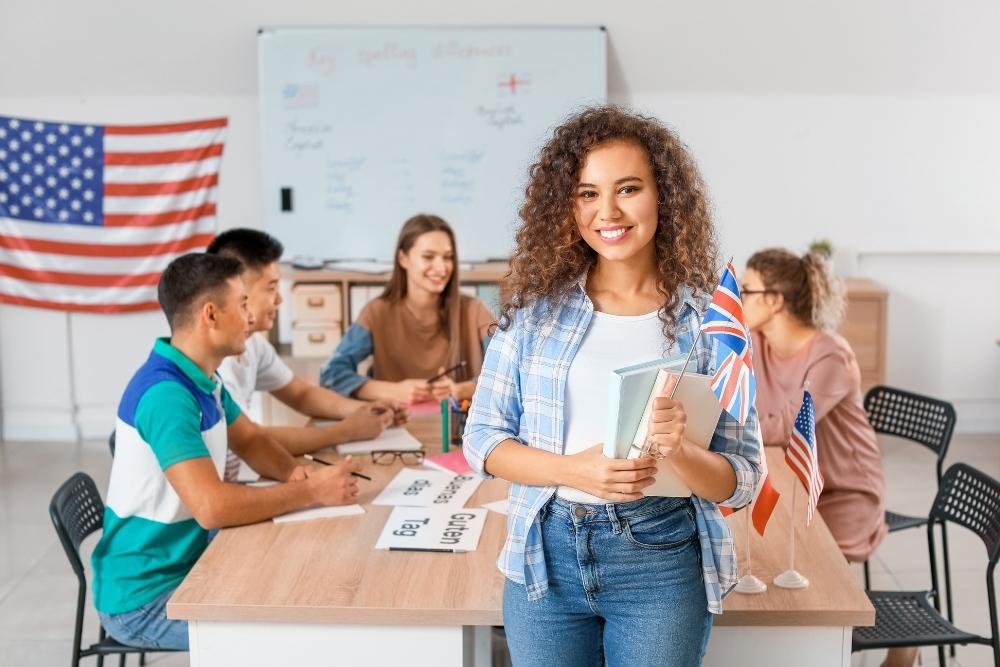 Uma jovem sorridente segura livros em uma sala de aula decorada com bandeiras de diferentes países. Ao fundo, colegas discutem em grupo, simbolizando um ambiente de aprendizado multicultural.