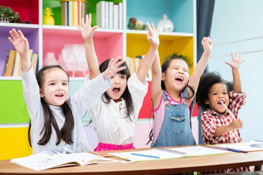 Crianças sorrindo e levantando as mãos em uma sala colorida.
