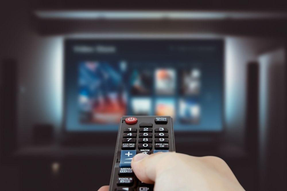 Pessoa segurando um controle remoto em direção a uma televisão com a interface de uma plataforma de streaming exibida na tela.