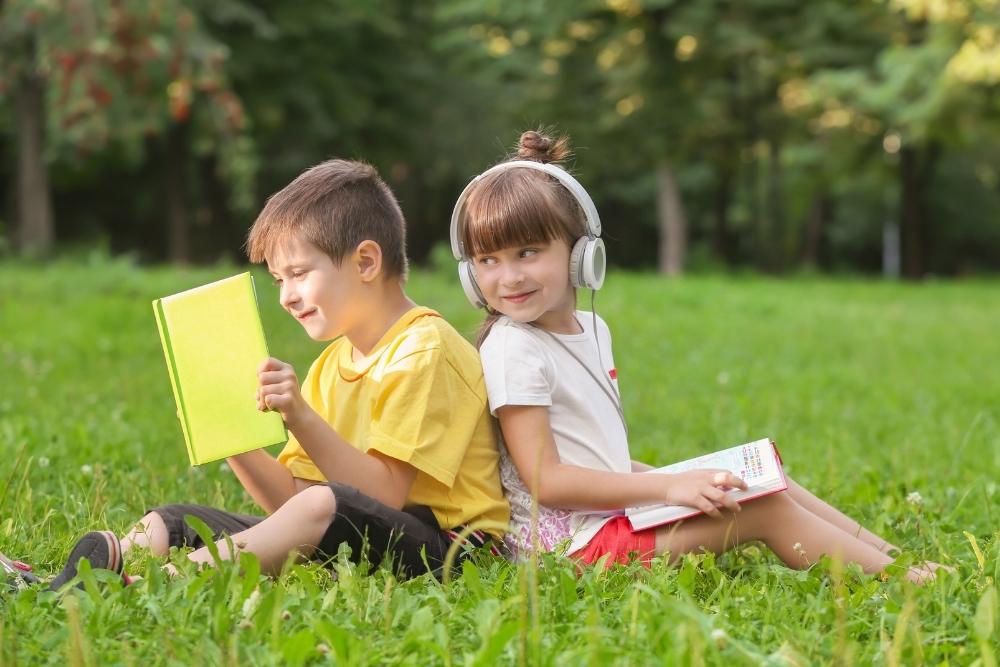 Duas crianças sentadas em um gramado, uma menina e um menino, ambos com livros coloridos e usando fones de ouvido, parecendo estar aproveitando um momento de leitura ao ar livre.