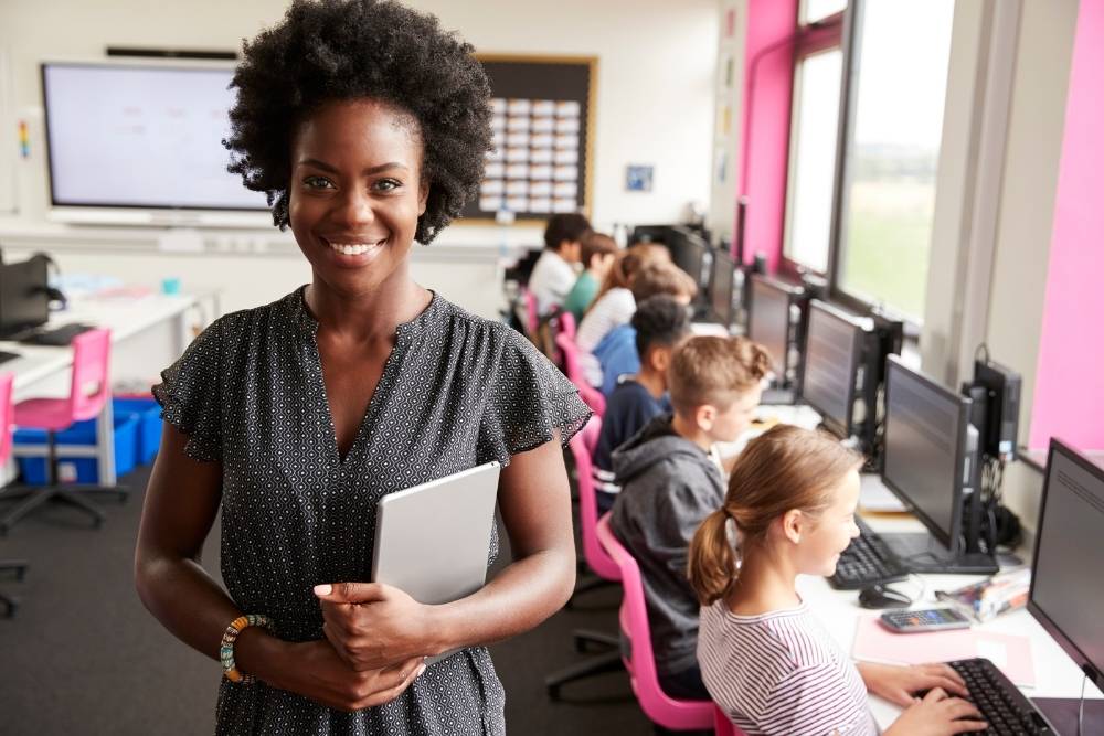 Professora sorridente em sala de aula com alunos em computadores.