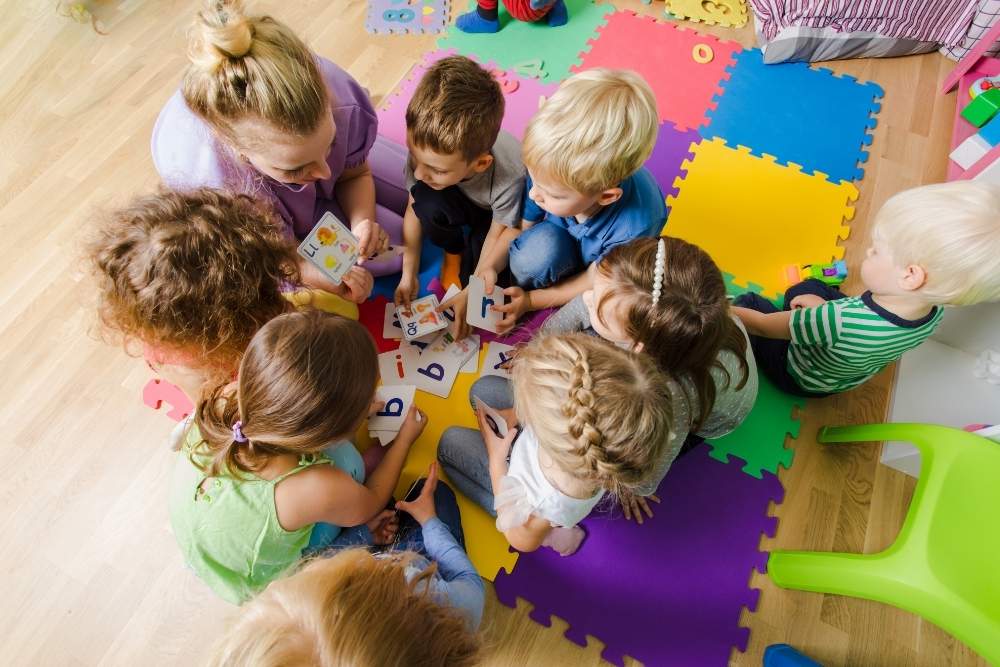 Grupo de crianças sentadas em um tapete colorido acompanhadas de uma educadora.