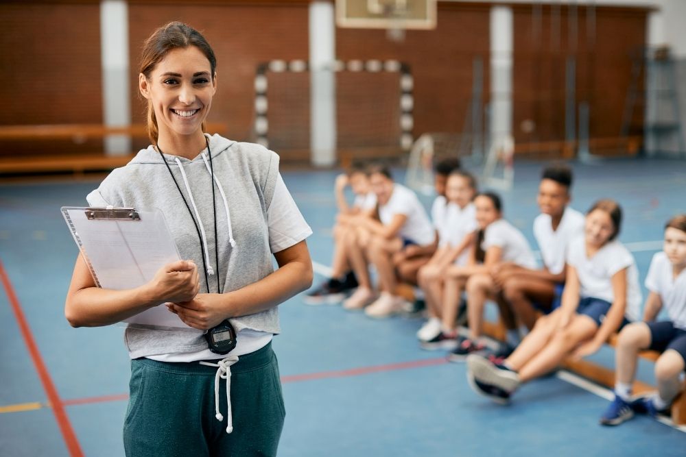 professora sorrindo em uma aula de Educação Física com alunos ao fundo.