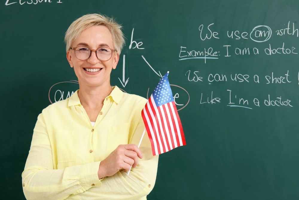 professora sorridente segurando uma bandeirinha dos Estados Unidos de costas para um quadro negro com palavras escritas em inglês.
